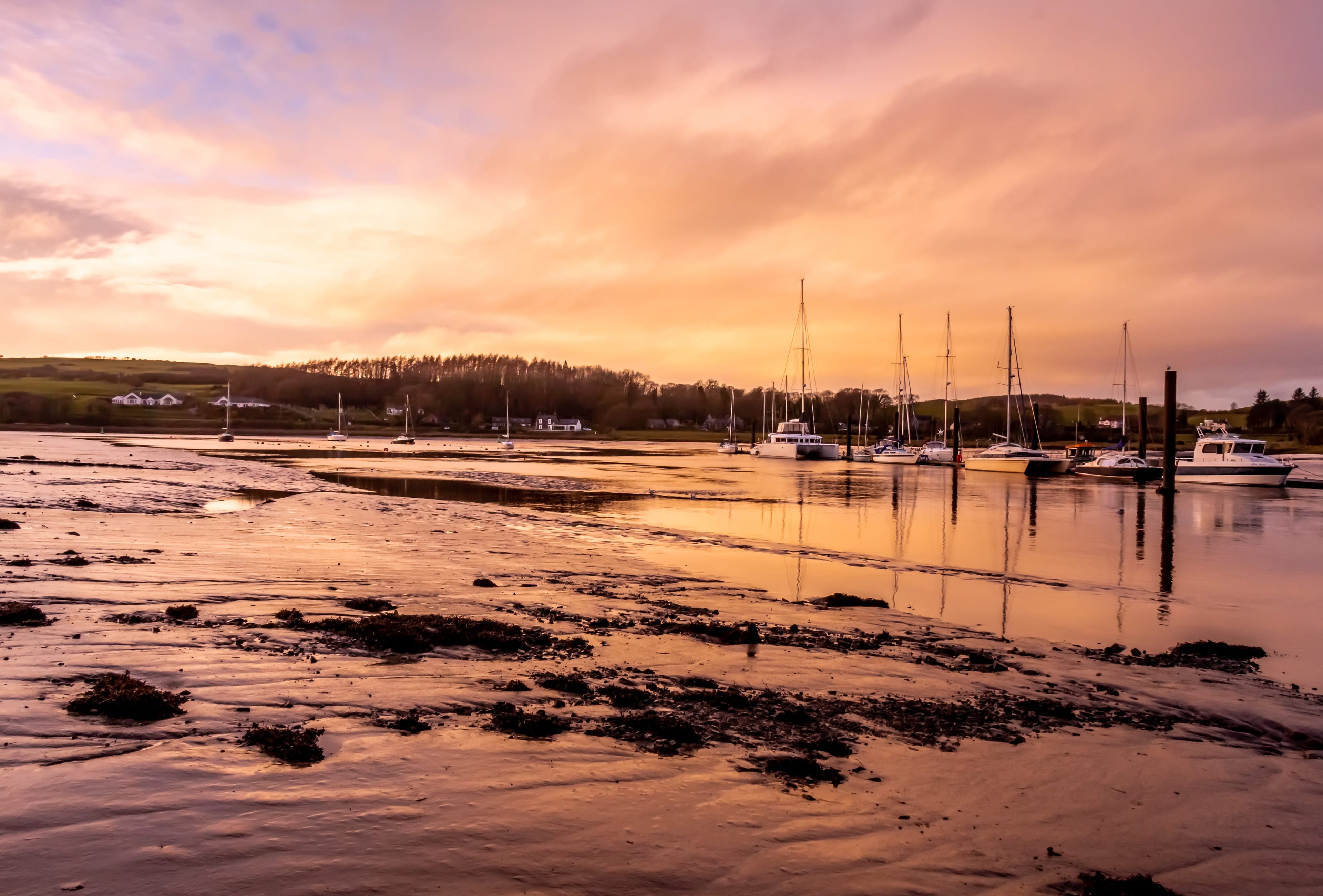Yachts and small boats moored on a tidal estuary at low tide, the wet sand and shallow channels turned pink and gold by a sunset, with farmland and a wooded ridge on the far shore